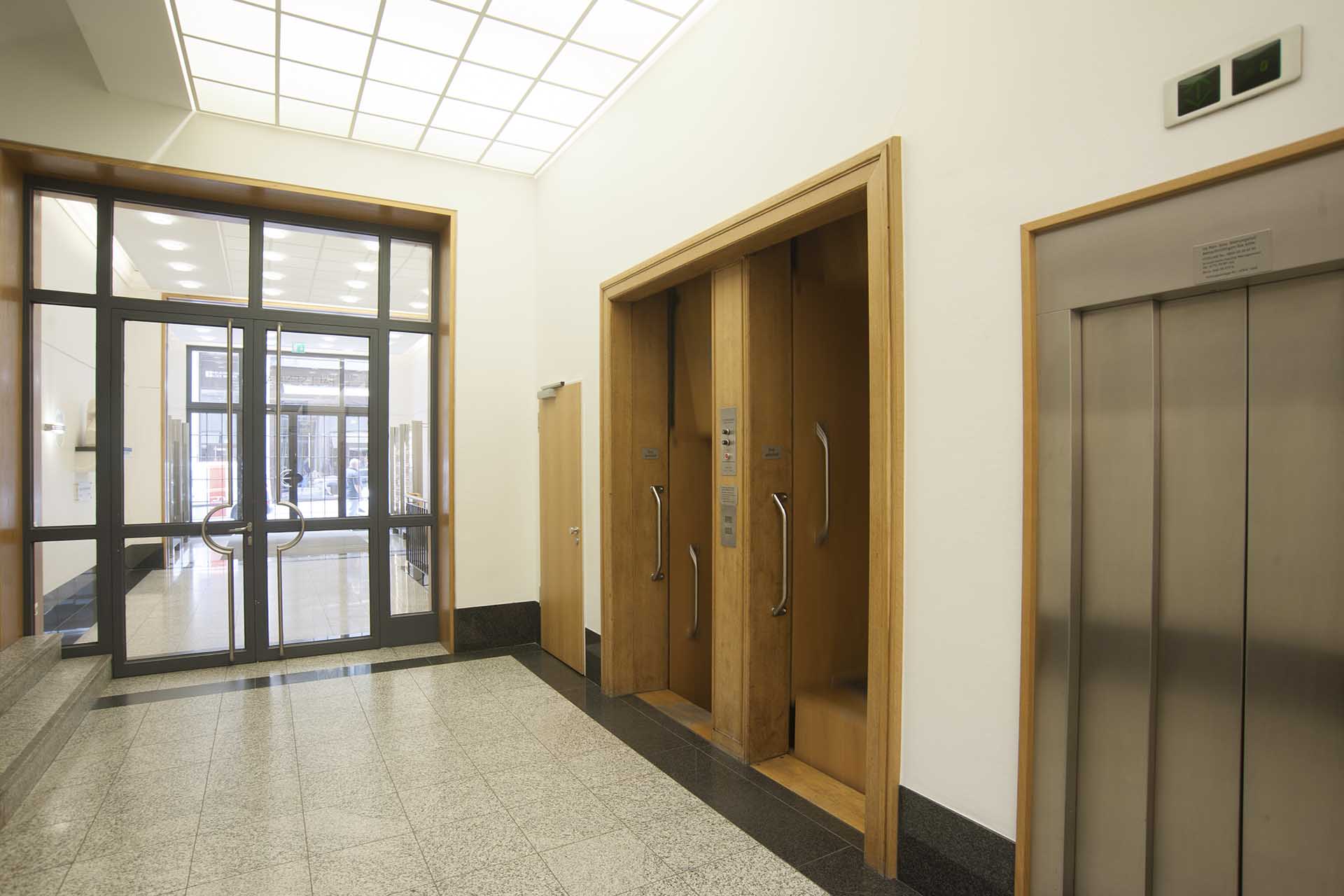 Foyer mit Glastür, holzverkleideten Durchgängen und Aufzug